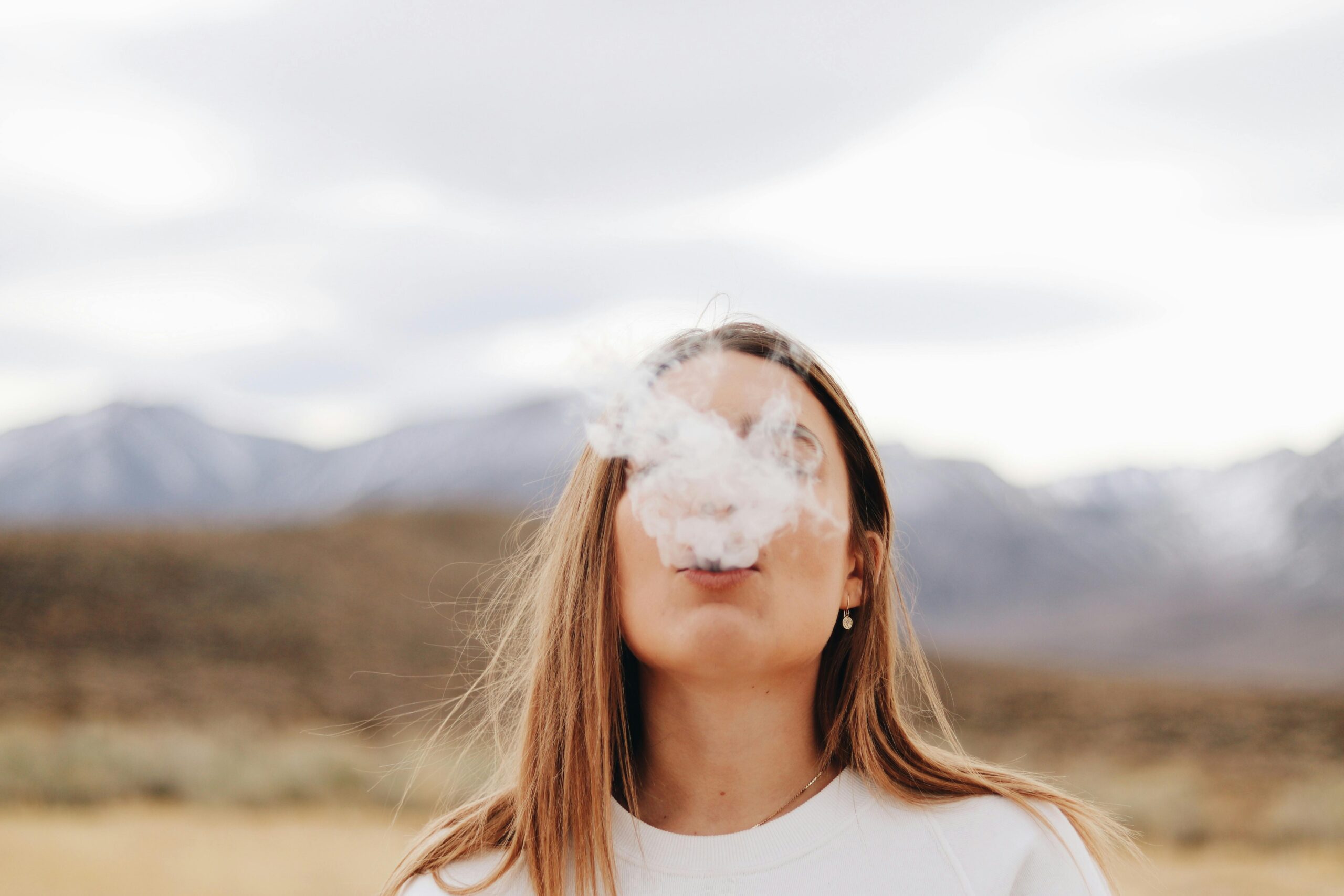 Woman with long hair exhaling smoke in an outdoor setting with mountains in the background.